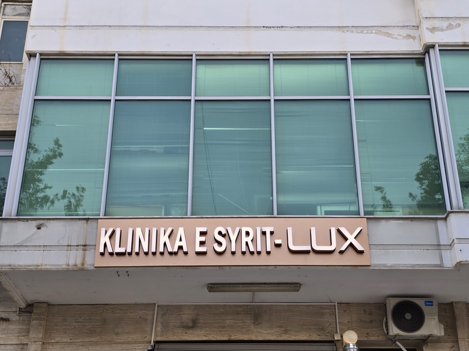 Shkronja 3D ngjyrë bakri KLINIKA E SYRIT-LUX mbi panel bezhë, nën fasadën me xham jeshil