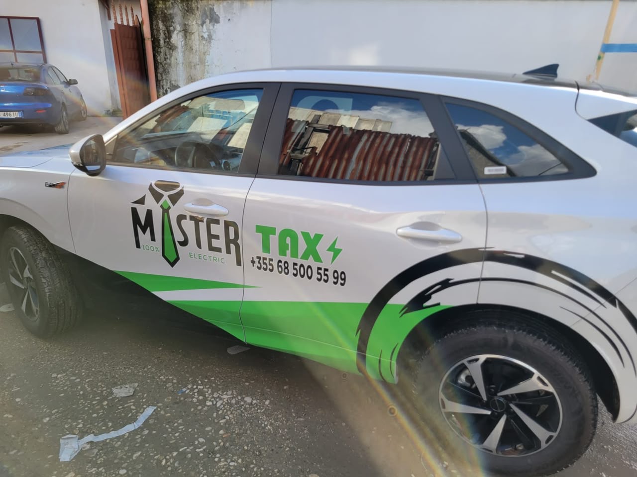 Taksi elektrike me grafika jeshile e të zeza Mister Taxi, logo kravate dhe numër telefoni në derë