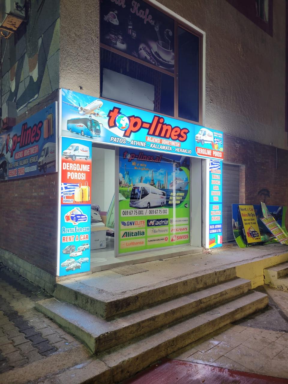 Fasadë qoshku e agjencisë Top-Lines me lightbox-e të ndezura dhe grafika vitrine natën