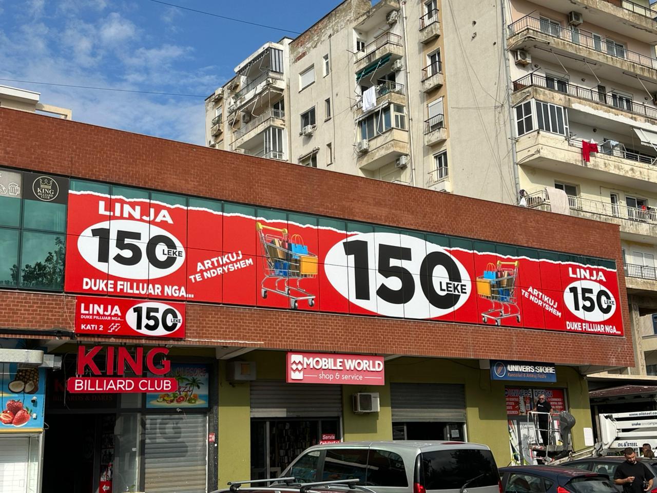 Panele të kuqe Linja 150 Lekë me karroca blerjesh përgjatë fasadës me tulla, mbi tabelat e King Billiard Club dhe Mobile World