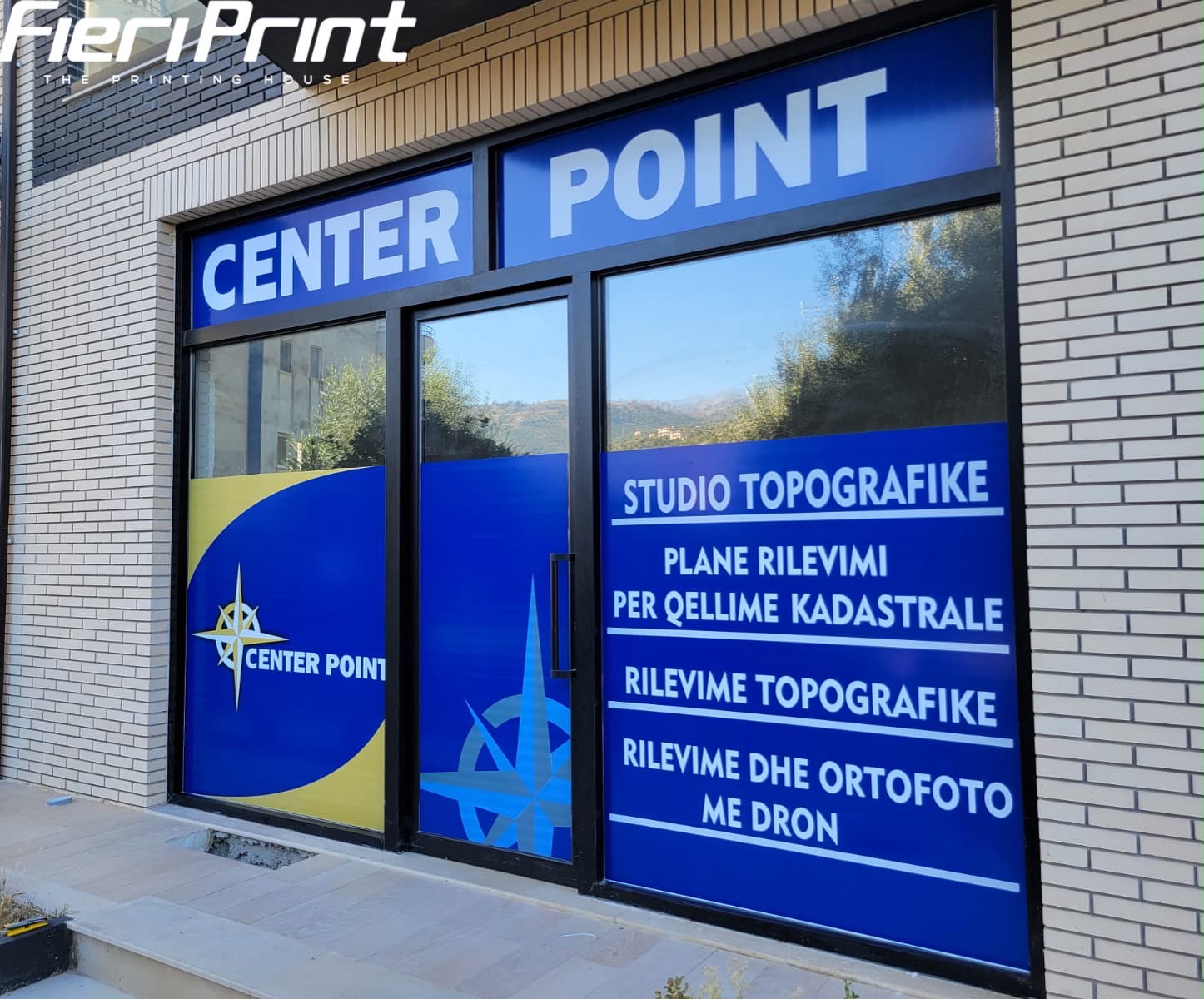 Vitrinë e Center Point me grafika blu e të verdha, logon me busull dhe listën e shërbimeve topografike
